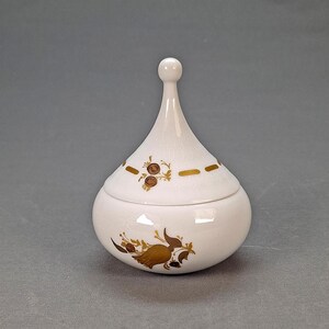 May include: A white ceramic jar with a lid, featuring gold floral and geometric designs. The jar has a bulbous base and a tall, slender lid with a rounded knob. The gold accents provide a contrast against the white background.