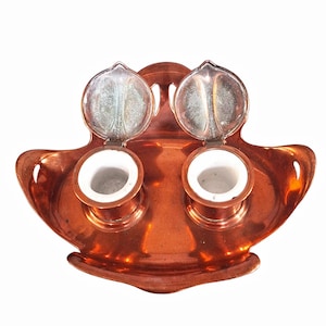 May include: Antique copper inkwell with two ceramic ink pots and hinged silver lids. The inkwell has a decorative, curved design with a polished finish. The lids are open, revealing the interior of the ink pots.