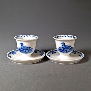 Op de afbeelding: Twee witte porseleinen eierdopjes met bijpassende schoteltjes. Elke beker heeft een blauw eendenontwerp en een blauwe bloemenrand. De schoteltjes hebben ook een blauw bloemmotief langs de randen. De set wordt gepresenteerd tegen een grijze achtergrond.