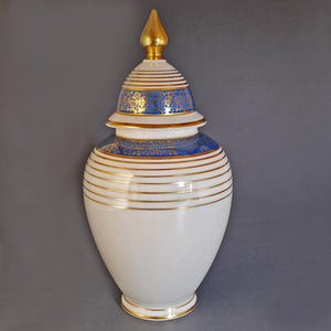 May include: A white ceramic jar with a gold lid and blue and gold decorative accents. The jar has a round body and a tall, narrow neck. The lid is topped with a gold finial.