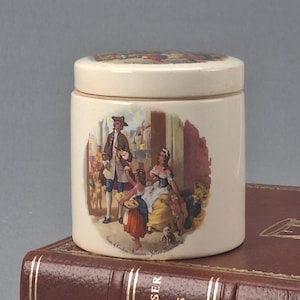 May include: A white ceramic jar with a lid featuring a colorful illustration of people in a street scene. The scene includes a man in a blue and gold suit, a woman in a green dress, and two children. The jar has the text "Gus A London Miniature - Yellow Dwarf" printed on the bottom.