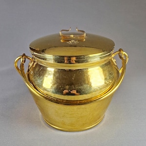 Puede incluir: Un recipiente de metal dorado con tapa y dos asas. El recipiente tiene una textura martillada y un acabado brillante. La tapa tiene un pequeño asa. El recipiente se utiliza probablemente para almacenamiento o exhibición.