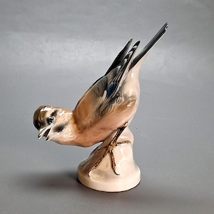 Puede incluir: Una figura de pájaro de cerámica en una pose dinámica, posada sobre una pequeña base de color claro. El pájaro presenta plumas detalladas en tonos de marrón, gris y azul, con el pico ligeramente abierto. La figura es un artículo decorativo.