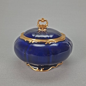 May include: A small, round, blue porcelain trinket box with a gold lid and trim. The lid has a decorative knob in the shape of a crown.