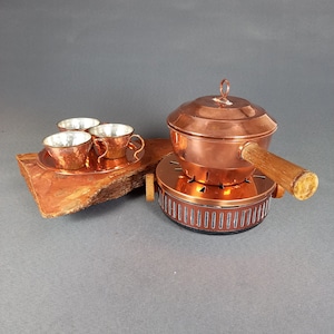 Puede incluir: Un juego de fondue de cobre con un mango de madera y tres pequeñas tazas de cobre sobre una base de madera.