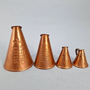 Puede incluir: Conjunto de cuatro tazas medidoras de cobre con marcas graduadas. Las tazas están anidadas, con la taza más grande en la parte inferior y la taza más pequeña en la parte superior. Las tazas son todas del mismo estilo, con un borde acampanado y un fondo redondeado.