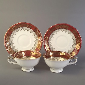 May include: Two white teacups and saucers with a red and gold ornate design. The rims of the cups and saucers are red with gold filigree. The cups have a gold interior and a small handle.