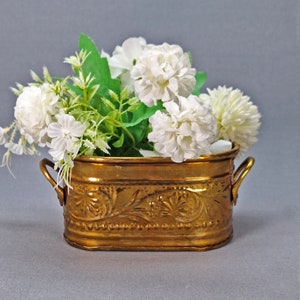 Puede incluir: Un macetero de latón con detalles ornamentados y dos asas, lleno de flores blancas y follaje verde.