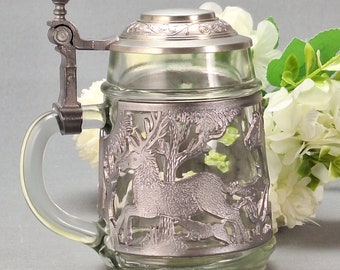 May include: A clear glass beer stein with a silver-colored metal lid and handle. The stein features a detailed relief of a deer in a forest scene. The handle and lid are also silver-colored.
