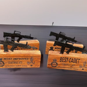Puede incluir: Cuatro trofeos de madera con modelos de rifles negros. Las bases de madera están grabadas con "MOST IMPROVED CORNWALL ACF SPRING CAMP 2024" y "BEST CADET CORNWALL ACF SPRING CAMP 2024".