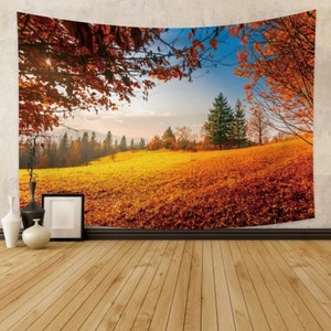 Op de afbeelding: Een wandkleed met een schilderachtig herfstlandschap met een veld van gouden bladeren, een bos op de achtergrond en een blauwe lucht met witte wolken. De bomen op de voorgrond hebben rode en oranje bladeren.