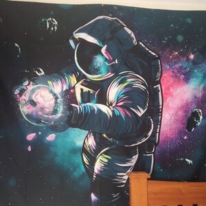May include: A colourful tapestry depicting an astronaut holding a glowing orb against a galaxy backdrop. The astronaut is wearing a white and blue spacesuit with a black helmet. The galaxy is a mixture of pink, purple, and blue colours.