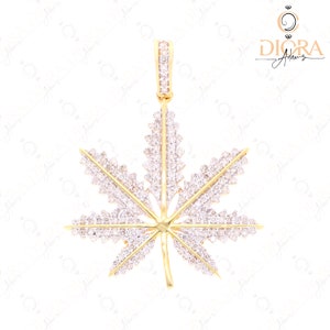 May include: A gold-colored pendant in the shape of a cannabis leaf, encrusted with numerous small, sparkling white gemstones. The pendant has a bail at the top for a chain. The design is intricate and detailed, with a focus on the leaf's shape.