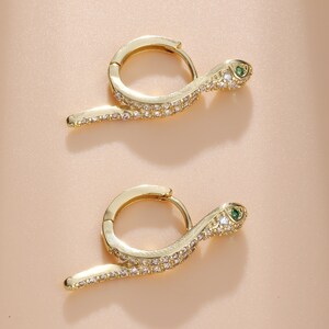 May include: Two gold-coloured hoop earrings, each shaped like a snake. The snake's body is set with small, clear crystals, and the head has a green gemstone eye. The earrings are shown against a light peach background.