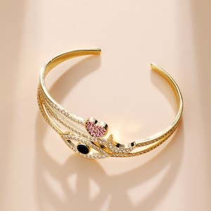 Peut inclure: Bracelet manchette doré avec un design décoratif. Le bracelet présente un accent en forme de cœur avec des pierres roses et un motif en forme d'œil avec des pierres claires et un centre noir. Le bracelet est ouvert en haut.