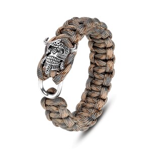May include: Braided paracord bracelet in shades of brown, gray, and tan. The bracelet features a detailed silver-colored skull charm with horns and a ring clasp. A rugged and stylish accessory.