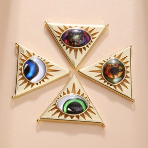 Puede incluir: Cuatro colgantes triangulares dorados con diseños de ojos. Cada colgante presenta un color de ojo diferente: azul, verde, marrón y un patrón de galaxia. Los triángulos tienen un diseño de sol alrededor del ojo.