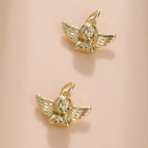 Pode incluir: Dois pingentes dourados em forma de anjo, com asas detalhadas e um halo. Os pingentes são suspensos por um pequeno laço, prontos para serem adicionados a um colar ou pulseira. O fundo é de uma cor pêssego suave.