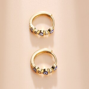May include: A pair of gold-coloured hoop earrings. Each earring has a series of small, blue and white evil eye charms and clear, round accents. The earrings are set against a soft, peach-coloured background.