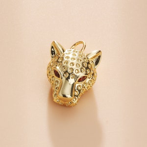 Puede incluir: Un colgante dorado con forma de cabeza de leopardo. El colgante tiene ojos rojos y una superficie texturizada con pequeños detalles redondos. La cabeza del leopardo está orientada hacia delante, con sus orejas y nariz claramente visibles.