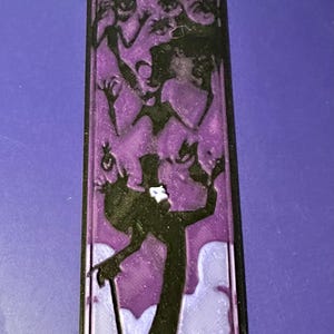 Puede incluir: Una pieza decorativa rectangular negra y morada que presenta una silueta de un hombre con bast&oacute;n y sombrero de copa, rodeado de otras figuras. El fondo es un degradado de morado y blanco.