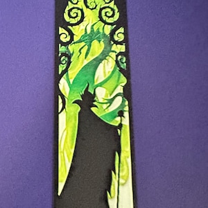Puede incluir: Un marcap&aacute;ginas rectangular negro con una ilustraci&oacute;n verde y amarilla de un personaje con un dise&ntilde;o de drag&oacute;n. El dise&ntilde;o tiene un efecto de vidriera con patrones arremolinados.
