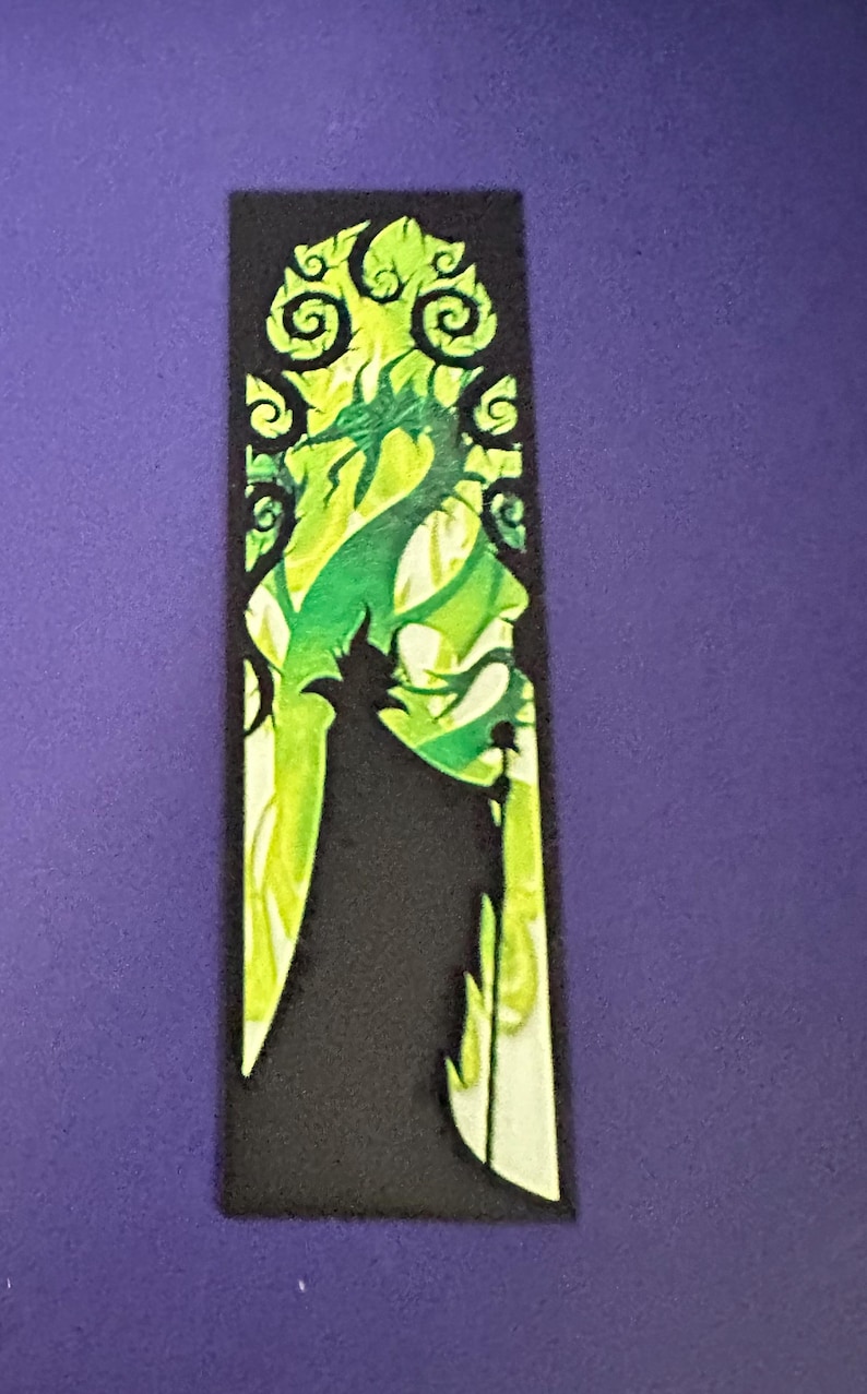 Puede incluir: Un marcap&aacute;ginas rectangular negro con una ilustraci&oacute;n verde y amarilla de un personaje con un dise&ntilde;o de drag&oacute;n. El dise&ntilde;o tiene un efecto de vidriera con patrones arremolinados.