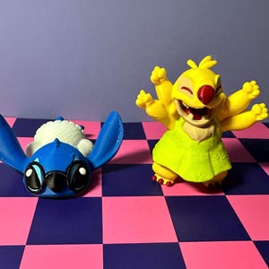 May include: Two character figurines. One is blue with large ears and eyes, lying on its back. The other is yellow with multiple arms, a red nose, and a green dress, posed with arms raised. They are on a checkered pink and blue surface.
