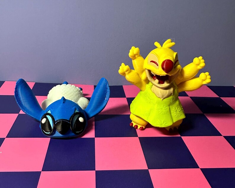 May include: Two character figurines. One is blue with large ears and eyes, lying on its back. The other is yellow with multiple arms, a red nose, and a green dress, posed with arms raised. They are on a checkered pink and blue surface.
