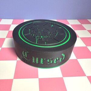May include: A black, round box with a green trim and the word "Cursed" in green lettering. The top of the box features a green outline of a skull and crossbones design. The box is sitting on a pink and white checkered surface.