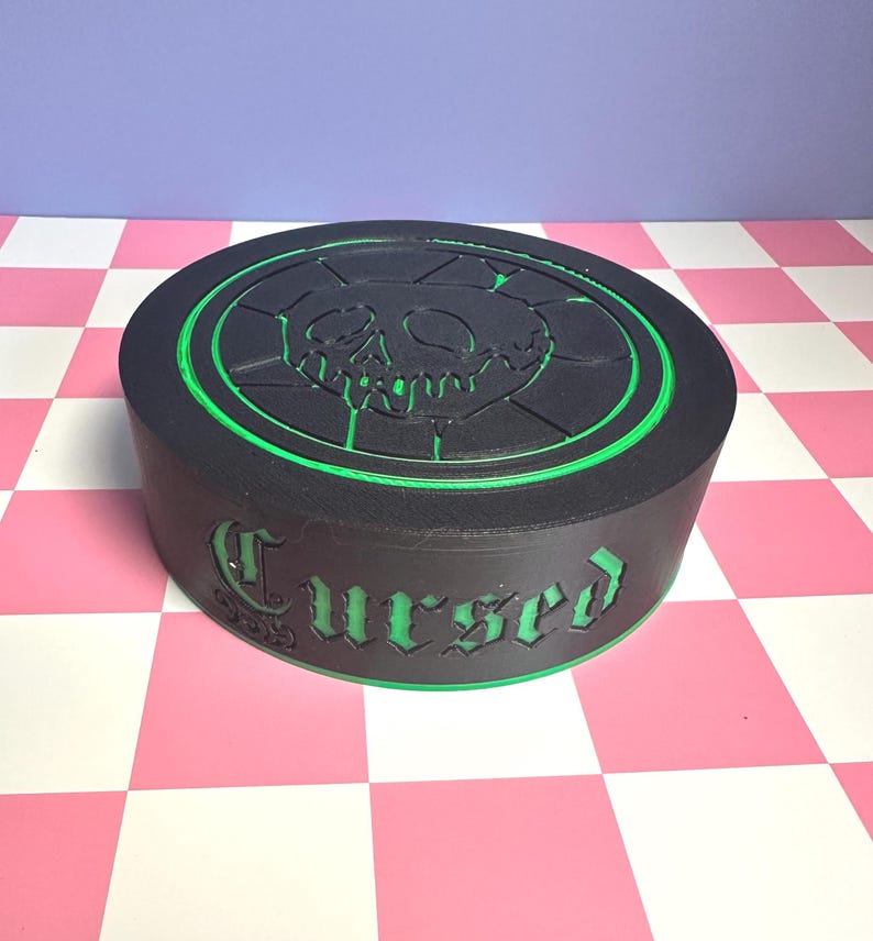 May include: A black, round box with a green trim and the word "Cursed" in green lettering. The top of the box features a green outline of a skull and crossbones design. The box is sitting on a pink and white checkered surface.