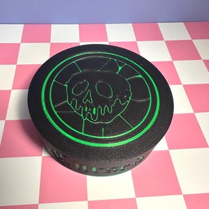 May include: Black, round box with a green skull and crossbones design. The skull is in the center, surrounded by a green circular border. The box has a textured surface and is sitting on a pink and white checkered surface.