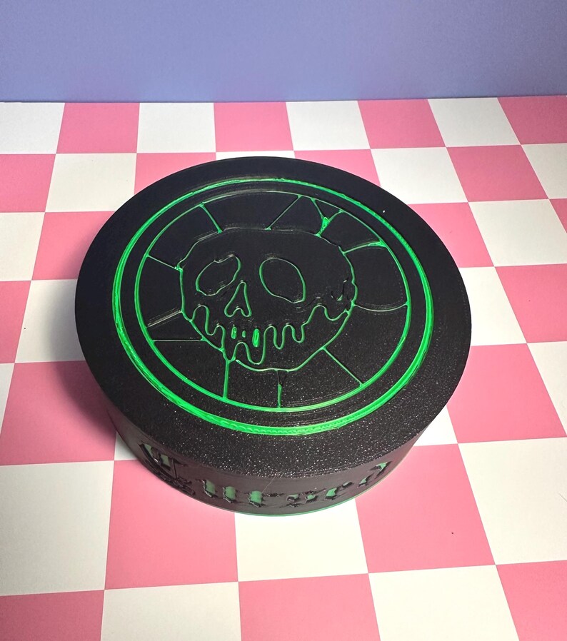 May include: Black, round box with a green skull and crossbones design. The skull is in the center, surrounded by a green circular border. The box has a textured surface and is sitting on a pink and white checkered surface.