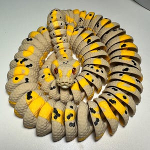 Puede incluir: Un juguete de serpiente articulado en espiral, con un cuerpo beige, detalles amarillos y manchas negras. La cabeza de la serpiente presenta ojos rojos y un dise&ntilde;o texturizado detallado. El juguete es de pl&aacute;stico y es flexible.