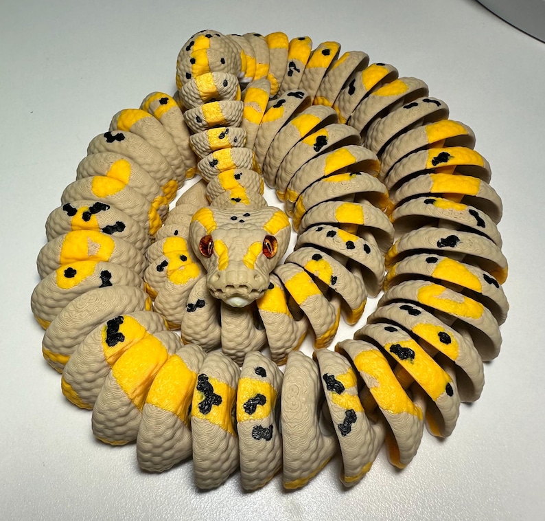 Puede incluir: Un juguete de serpiente articulado en espiral, con un cuerpo beige, detalles amarillos y manchas negras. La cabeza de la serpiente presenta ojos rojos y un dise&ntilde;o texturizado detallado. El juguete es de pl&aacute;stico y es flexible.