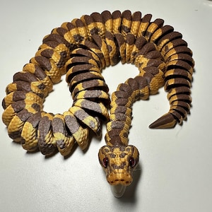 Puede incluir: Una serpiente de juguete articulada y detallada en tonos marrones y amarillos. El cuerpo de la serpiente est&aacute; segmentado, lo que permite un movimiento flexible. La cabeza presenta ojos rojos y una lengua b&iacute;fida, con un patr&oacute;n de escamas detallado.