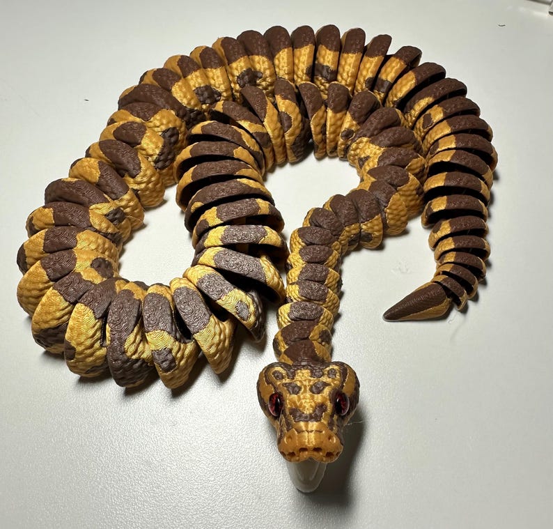 Puede incluir: Una serpiente de juguete articulada y detallada en tonos marrones y amarillos. El cuerpo de la serpiente est&aacute; segmentado, lo que permite un movimiento flexible. La cabeza presenta ojos rojos y una lengua b&iacute;fida, con un patr&oacute;n de escamas detallado.