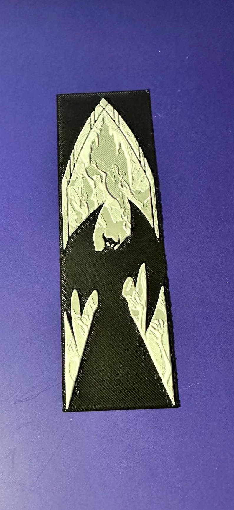 Puede incluir: Un emblema rectangular negro con un dise&ntilde;o plateado en forma de murci&eacute;lago. El emblema tiene una superficie texturizada y est&aacute; sobre un fondo p&uacute;rpura. El dise&ntilde;o del murci&eacute;lago est&aacute; detallado con un estilo arquitect&oacute;nico g&oacute;tico.