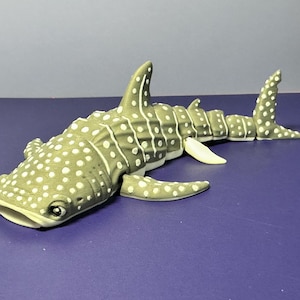 May include: A detailed figurine of a whale shark, featuring a gray body with white spots and fins. The model has a realistic design, with a detailed head, fins, and tail. The toy is set against a purple background.