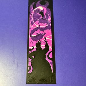 Puede incluir: Marcap&aacute;ginas negro con la silueta de un personaje con tent&aacute;culos, sobre un fondo rosa y morado con formas arremolinadas. El marcap&aacute;ginas es rectangular y tiene un aspecto texturizado.