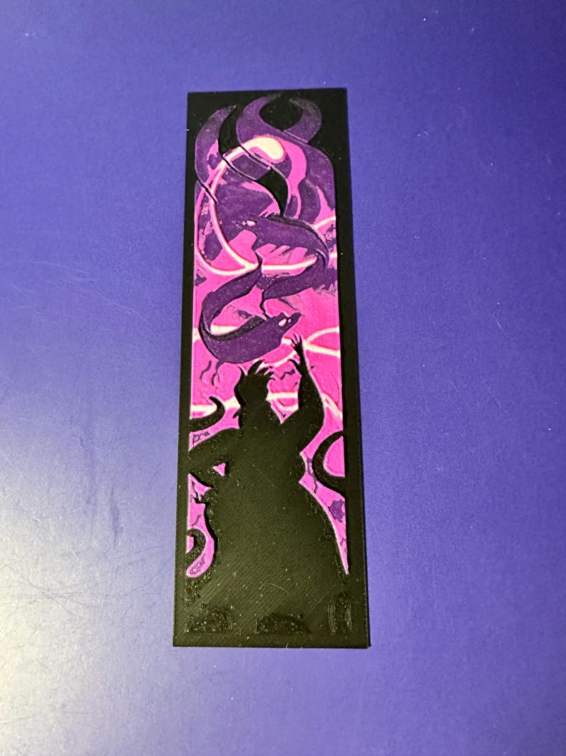 Puede incluir: Marcap&aacute;ginas negro con la silueta de un personaje con tent&aacute;culos, sobre un fondo rosa y morado con formas arremolinadas. El marcap&aacute;ginas es rectangular y tiene un aspecto texturizado.