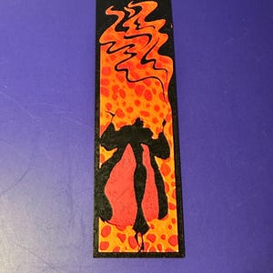 Puede incluir: Marcap&aacute;ginas rectangular con una ilustraci&oacute;n estilizada. El dise&ntilde;o presenta una silueta negra de una figura con una capa roja, sobre un fondo de llamas naranjas y amarillas con un patr&oacute;n de puntos. El marcap&aacute;ginas tiene un borde negro.