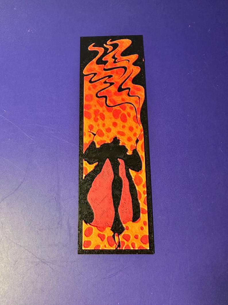 Puede incluir: Marcap&aacute;ginas rectangular con una ilustraci&oacute;n estilizada. El dise&ntilde;o presenta una silueta negra de una figura con una capa roja, sobre un fondo de llamas naranjas y amarillas con un patr&oacute;n de puntos. El marcap&aacute;ginas tiene un borde negro.