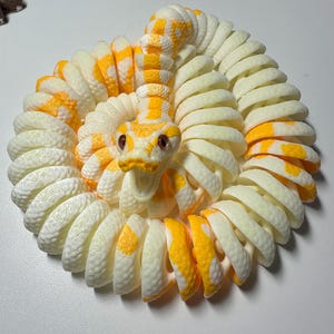 Puede incluir: Una figura de serpiente enroscada en crema y amarillo. El cuerpo de la serpiente es de color crema con marcas amarillas, y su cabeza es amarilla con ojos rojos. La serpiente est&aacute; enroscada en un patr&oacute;n circular, creando una pieza decorativa.