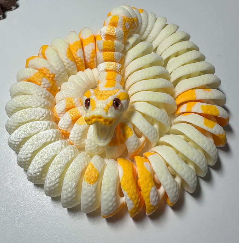 Puede incluir: Una figura de serpiente enroscada en crema y amarillo. El cuerpo de la serpiente es de color crema con marcas amarillas, y su cabeza es amarilla con ojos rojos. La serpiente est&aacute; enroscada en un patr&oacute;n circular, creando una pieza decorativa.