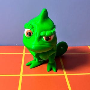 May include: A green chameleon figurine with a grumpy expression. The figurine has large, orange eyes and is posed on a red and yellow grid surface. The background is a solid blue color.