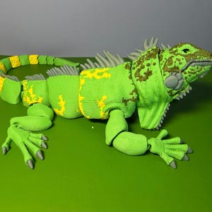 May include: A green articulated iguana figure with yellow patches along its back and tail. The lizard features a textured skin surface, grey spikes running along its spine, and grey claws on its feet. The head has brown mottled markings.