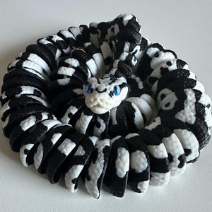 Puede incluir: Escultura de serpiente enroscada en blanco y negro, con un patr&oacute;n detallado. La cabeza de la serpiente est&aacute; en el centro, con ojos azules. La escultura est&aacute; hecha de un material que parece ser impreso en 3D.