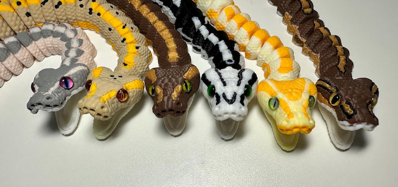 Puede incluir: Colecci&oacute;n de figuras de serpientes coloridas y detalladas. Las serpientes var&iacute;an en color, incluyendo gris, beige, marr&oacute;n, negro y amarillo, con patrones intrincados y ojos realistas. Cada serpiente est&aacute; curvada.