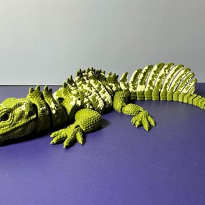 May include: A green and white articulated lizard toy, lying on a purple surface. The toy has a detailed, textured design, with visible scales and a segmented body. The lizard's head is facing left, and its legs are extended.